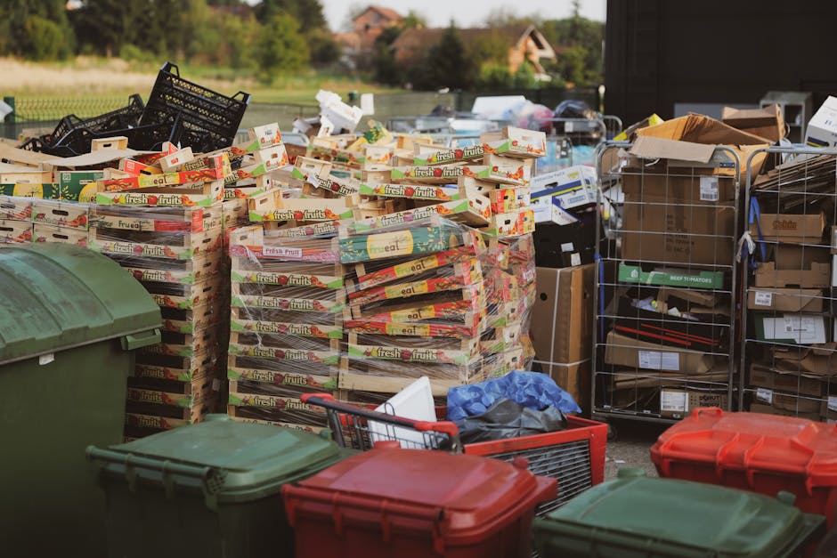 A large outdoor collection of various waste materials, including multiple stacks of empty cardboard boxes labeled with 'fresh fruits' on the sides, positioned on a paved surface in front of a dark bui