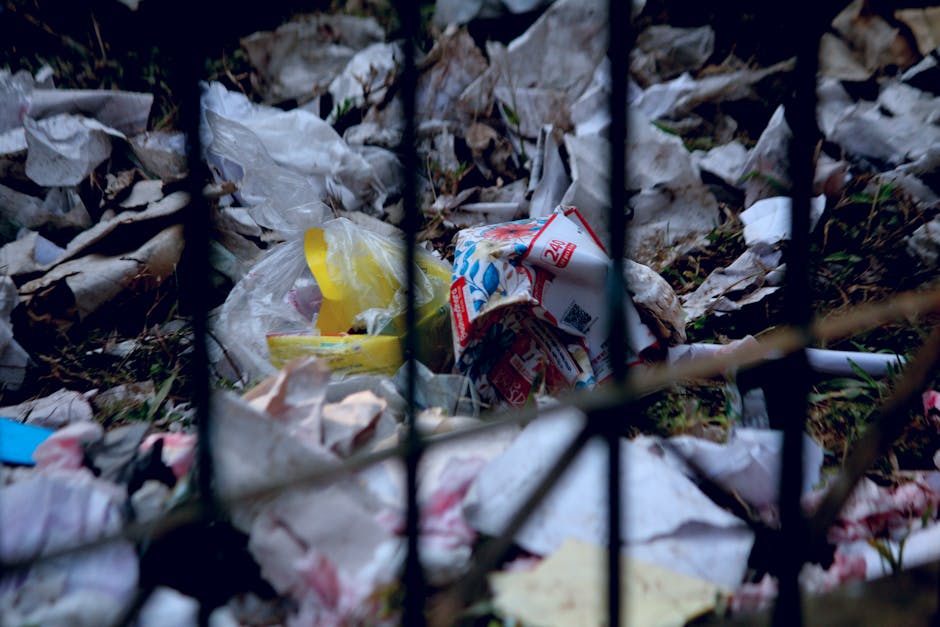 A collection of overflowing waste bins and scattered rubbish piled on a paved area near a parking space, with various types of waste including cardboard boxes, plastic bags, paper, and packaging mater