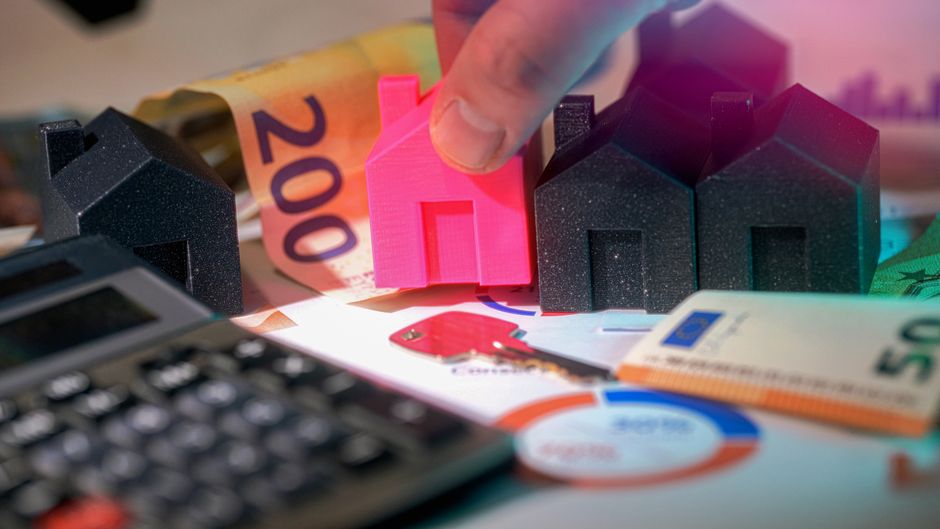 A close-up image showing a person's hand placing a small pink house-shaped object among three larger black house-shaped models, which are arranged on a cluttered surface that includes a calculator, eu