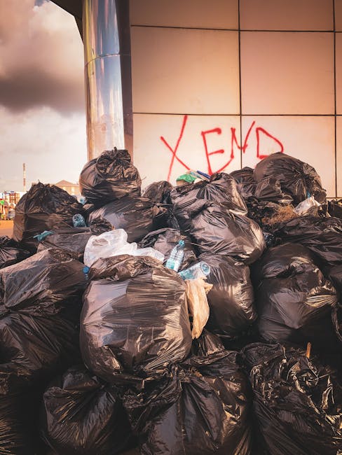 A large accumulation of black plastic rubbish bags stacked against a tiled urban wall, with some bags appearing overfilled and torn open, revealing plastic bottles and miscellaneous waste inside. The 