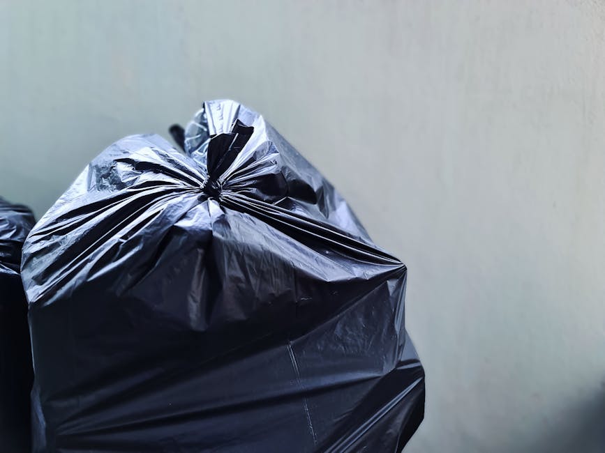 A large accumulation of black plastic rubbish bags stacked against a tiled urban wall, with some bags appearing overfilled and torn open, revealing plastic bottles and miscellaneous waste inside. The 